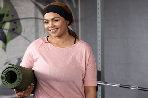 African american woman holding yoga mat and smiling in urban gym with squat rack
