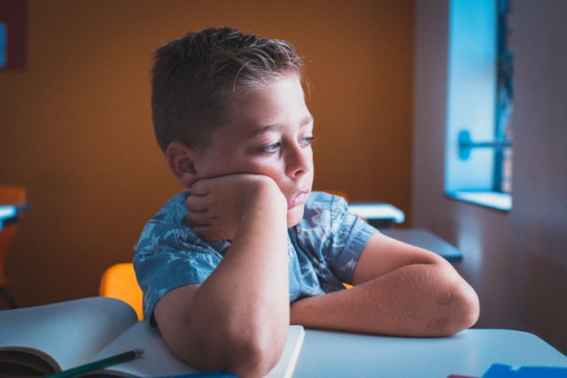 Young boy sitting at a school desk, appearing bored and daydreaming while looking out of the window. Ideal for use in educational materials, articles on childhood education, or content discussing student engagement and classroom dynamics.