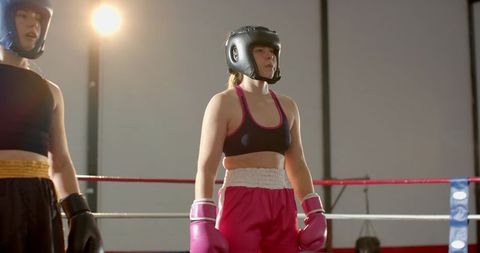 Female boxer standing in ring wearing pink gloves and headgear under spotlight