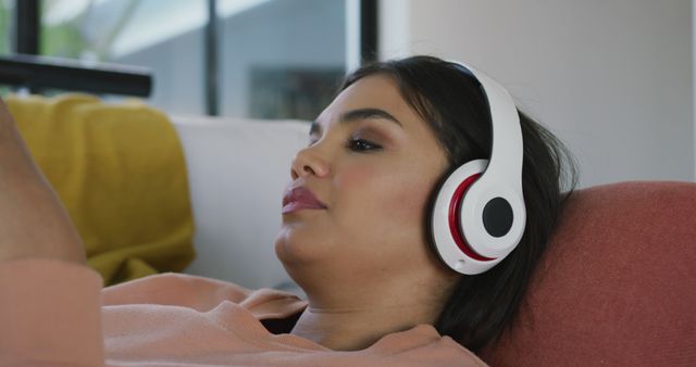 Image of relaxed biracial woman lying on sofa with headphones and listening to music. leisure, relax, spending free time with technology at home concept.