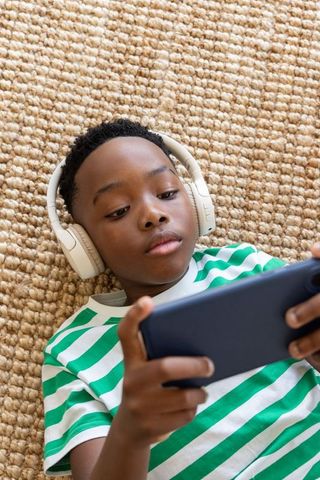African american boy lying on jute rug wearing headphones and green striped tee using phone