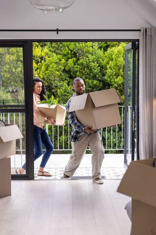 Diverse couple carrying moving boxes and potted plant into new home through glass doors