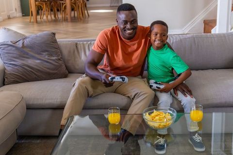 African American Father and Son Playing Video Games on Living Room Sofa with Snacks