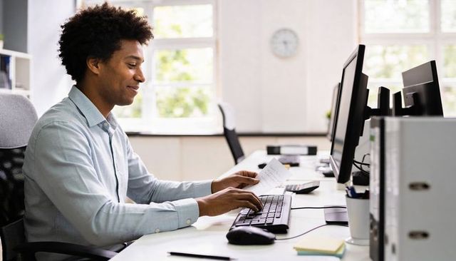 Young professional reviewing documents and typing at modern open-plan office workstation