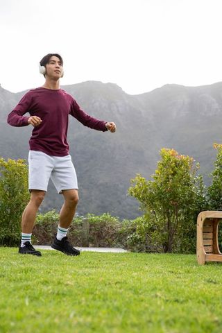 Asian man dancing on grassy lawn with headphones at mountain foothills enjoying outdoor fitness