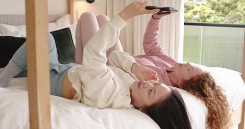 Young diverse women relaxing on bed using smartphone together in bright minimalist bedroom