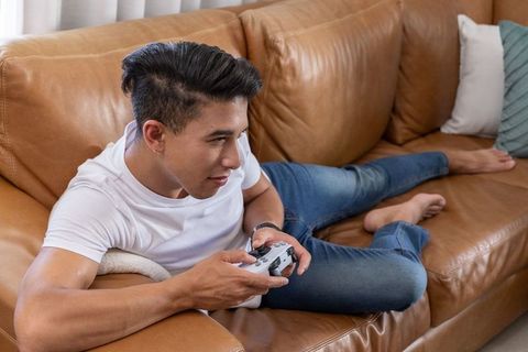 Asian man reclining on leather sofa playing video games with controller casual home leisure