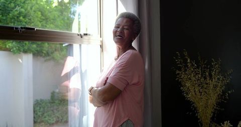 Senior woman leaning by window smiling folding arms in sunlit home with garden view
