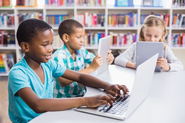 Children are engaged in using a laptop and tablets in a school library. This image is perfect for illustrating modern education, technology integration in classrooms, and diverse learning environments. It can be used in educational materials, school websites, and articles about digital learning.