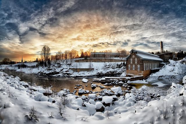 This picturesque scene captures a snow-covered riverbank with a charming abandoned brick building at sunset. The expansive sky is painted with soft, colorful clouds, adding warmth to the cold, serene landscape. Perfect for projects related to winter, travel, nature, and tranquility.