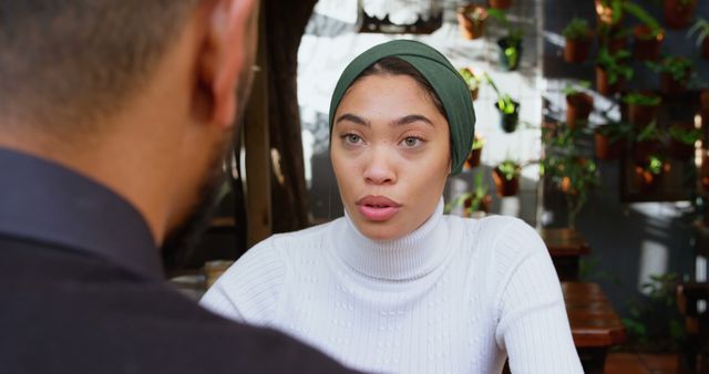 Woman wearing green headscarf and white sweater having a conversation with a friend in a cozy café with indoor plants on the background. Perfect for depicting themes of friendship, communication, discussing ideas, or interactions in social settings.