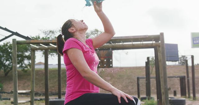 Tired caucasian woman in pink t shirt sitting and refreshing herself from water bottle at bootcamp. Female fitness, challenge and healthy lifestyle.