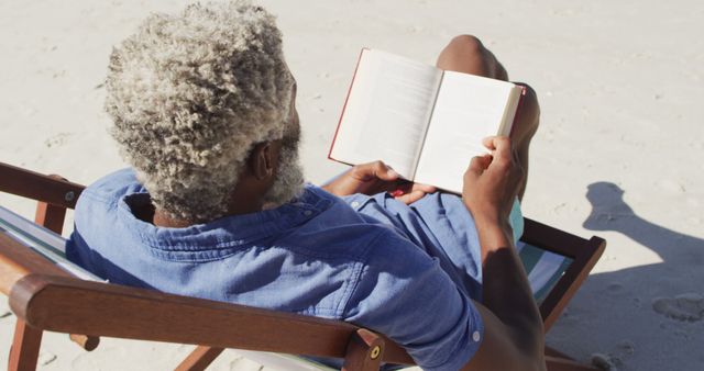 Elderly man with gray hair reading book while sitting in deck chair on beach. Scene is perfect for illustrating relaxation, retirement, leisure activities for seniors, vacation concepts, and serene outdoor settings.