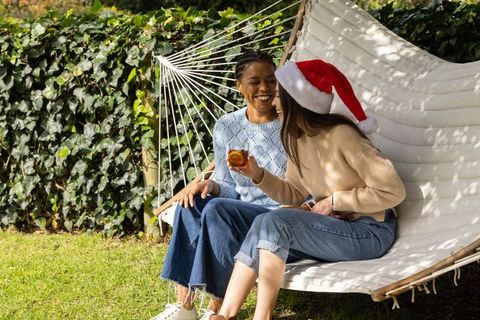 Friends relaxing on hammock wearing santa hat, sweaters and jeans, sharing drink outdoors