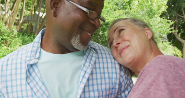 Senior interracial couple bonding outdoors, woman resting her head on man's shoulder while both share a happy moment. Ideal for promoting themes of love, retirement, joy, togetherness, and healthy relationships in senior years.