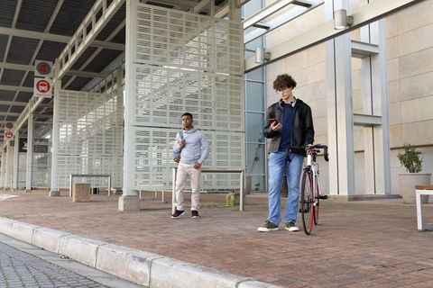 Young men waiting at curbside with bicycle and checking smartphone under transit canopy