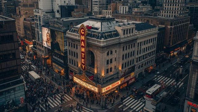 Historic theater marquee glowing over crowded urban intersection with neon signs and buses at dusk