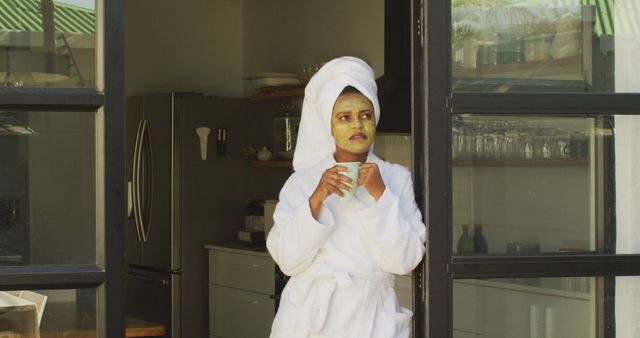 Happy african american woman with beauty mask on face, drinking coffee in kitchen. domestic lifestyle, spending free time at home.