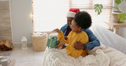Couple exchanging cozy christmas gift on couch, woman opening present, man wearing santa hat