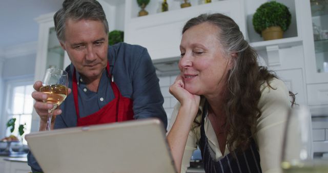 Happy senior caucasian couple in kitchen looking at laptop, talking and enjoying glass of white wine. retirement lifestyle together at home.