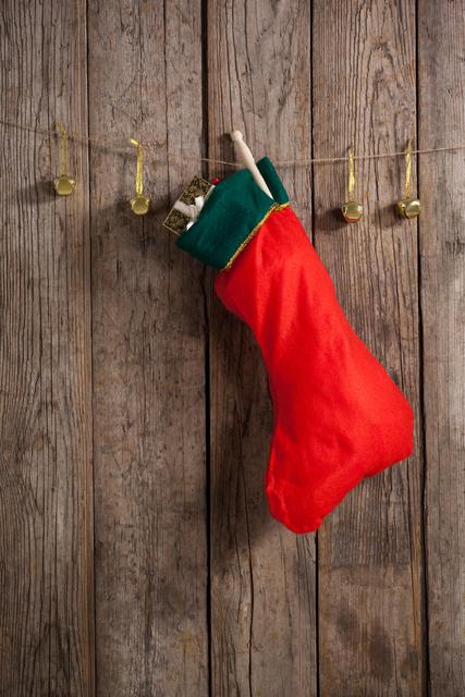 A red Christmas stocking filled with small gifts is hanging alongside small golden bells on a string against a rustic wooden wall. This scene evokes a traditional holiday atmosphere. Ideal for use in Christmas-themed promotions, holiday greeting cards, and festive home decor inspiration.