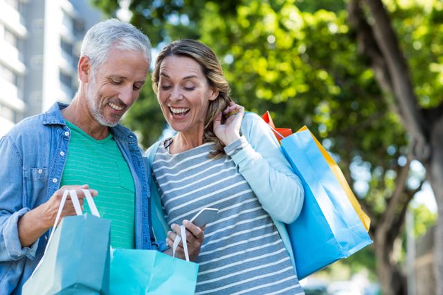 Happy mature couple using mobile phone while holding shopping bags in city