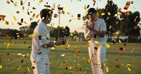 Cricket teammates celebrating victory with gold confetti falling on sunset grassy field