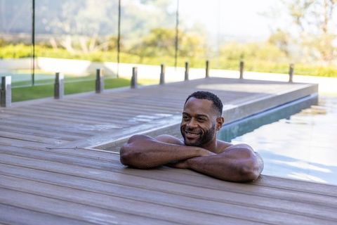 Mature african man relaxing and smiling at pool edge on wooden deck in sunlit backyard