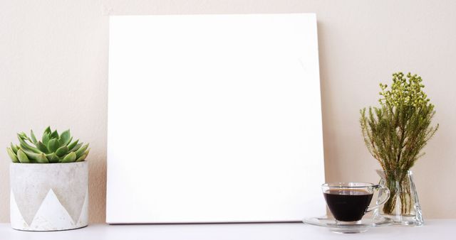 Blank canvas placed on white surface with a cup of coffee and plants arranged neatly. Ideal for promoting art supplies, organizing workspaces, or designing minimalist home office settings. Great for use in advertisements, blogs, and social media promoting creative inspiration, interior decor, and lifestyle aesthetics.