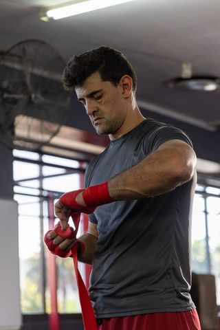 Focused boxer wrapping red hand wraps before training in sunlit gym wearing gray top
