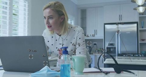 Woman working from home in modern kitchen leaning over laptop with headset and face mask