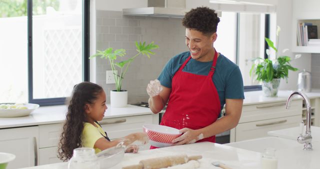 This image depicts a joyful moment where a biracial father and his young daughter are baking together in a modern kitchen. They both appear to be smiling, embodying the warmth and affection in their family bond. Ideal for use in content related to family activities, parenting tips, home life inspiration, promoting cooking classes, or advertisements targeting family-oriented products. This image promotes positive family interactions and showcases the importance of spending quality time together.