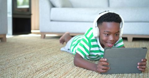 African american boy lying on rug playing with tablet wearing headphones relaxing at home