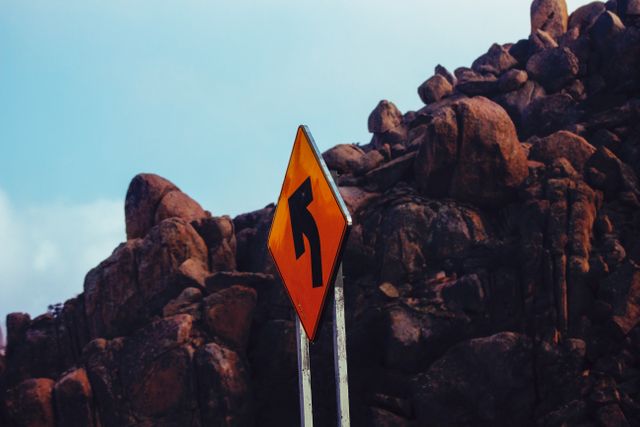 Orange road sign indicating a left curve stands tall amidst a rocky mountain terrain. Excellent for use in travel articles, road safety campaigns, or transportation blogs, emphasizing navigation through rugged landscapes. The striking contrast between the bright sign and the rugged rocks makes it visually appealing for travel advertisements or guidebooks.