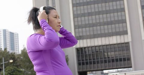Mature woman adjusting white headphones in urban plaza wearing purple activewear for workout