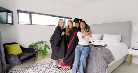Multiracial friends celebrating graduation taking selfie in modern bedroom with gowns