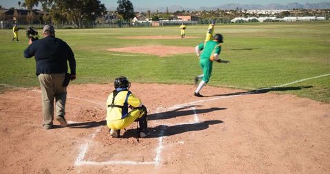 Youth baseball catcher squatting at home plate while runner rounding base during sunny game