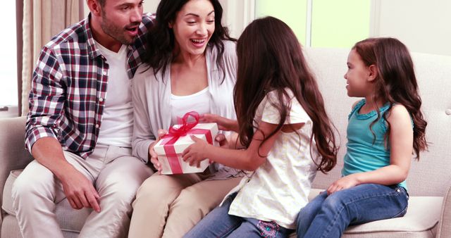 Family enjoying a joyful moment while exchanging gifts in a cozy living room. Perfect for advertisements showcasing family bonds, holiday celebrations, or promoting home and lifestyle products. Use to illustrate themes of love, happiness, and togetherness.