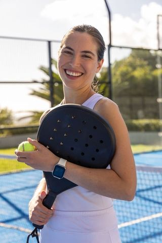 Smiling woman holding padel racket and tennis ball on blue outdoor court with smartwatch