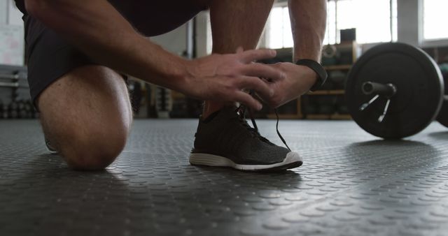 Male athlete tying shoe lace on gym floor, getting ready for workout, barbell in background. Useful for fitness blogs, exercise preparation tutorials, sportswear promotions.