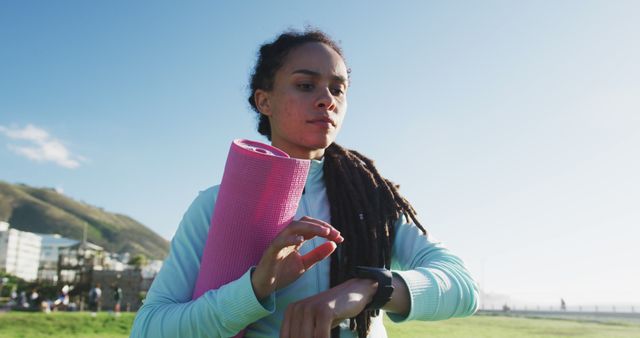 Young woman with dreadlocks holding pink yoga mat while checking fitness tracker in grassy field on a sunny day. Ideal for content related to fitness, health, wellness, outdoor activities, and new year fitness resolutions.