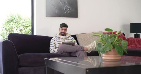 Indian man relaxing on dark velvet sofa using tablet in modern minimalist living room