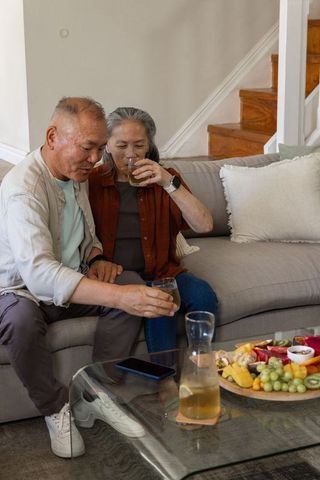 Mature asian couple sipping drinks and reaching for glasses on cozy living room sofa