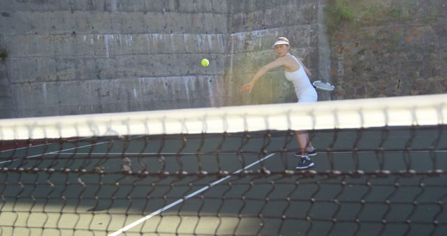 Woman serving tennis ball on outdoor court on sunny day. Visible warm weather adds to sporty and active vibe. Composition suitable for use in advertisements for sports equipment, fitness programs, or wellness and athletic campaigns. Showcases energy, dedication, and athleticism.