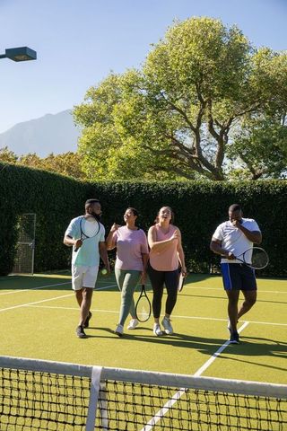 Diverse friends walking on tennis court carrying rackets on sunny day