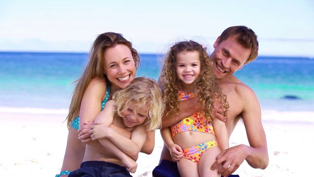 This depicts a joyful family of four embracing on a sunny beach. With turquoise water in the background, this could be used in advertisements or promotions for family vacations, travel brochures, or wellness campaigns focusing on family bonding time.