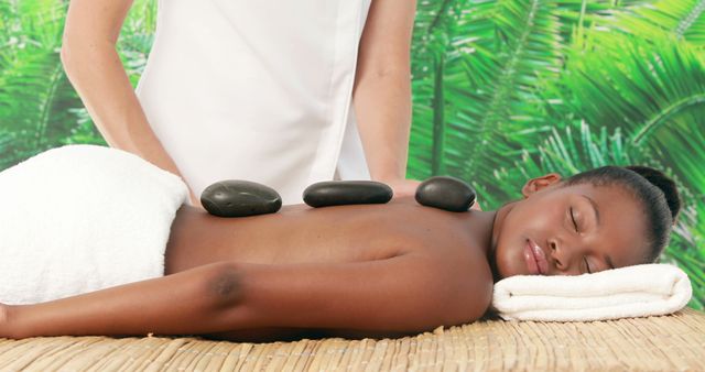An African American woman enjoys a relaxing hot stone massage at a spa, with copy space. Her serene expression and the tropical backdrop suggest a tranquil and rejuvenating experience.