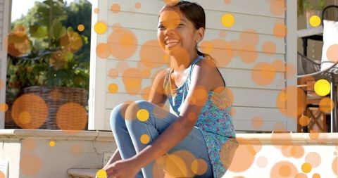 Smiling girl sitting on porch stoop wearing floral top blue leggings with sunlit bokeh