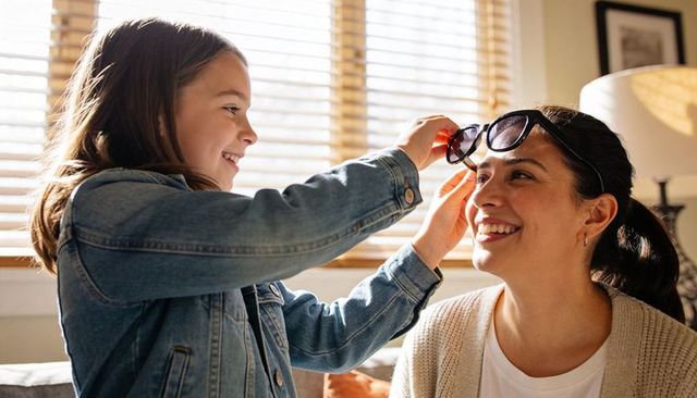 Daughter placing sunglasses on smiling mother in sunlit cozy living room moment