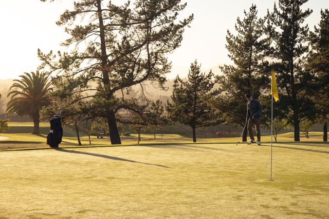 Mid distance view of caucasian young man hitting golf ball towards hole with yellow flag. Tree, golf course, sunlight, golf, unaltered, nature, sport and weekend activities concept.